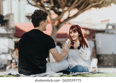 Two friends share a conversation while sitting on a blanket outdoors under the sunlight, enjoying each other's company and a relaxing time.