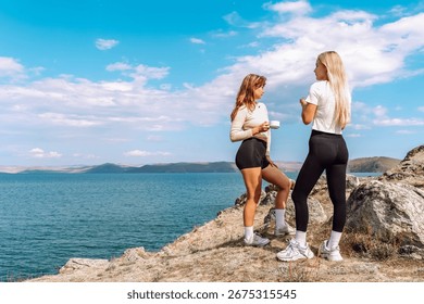 Two friends share tea while enjoying the stunning lakeside view under a bright sky.