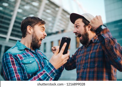 Two Friends Showing Sincere Emotions Of Joy About Victory In Online Lottery. Men Being Happy Winning A Bet In Online Sport Gambling Application With Football Stadium On The Background. 