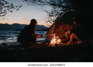 Two friends sit by a campfire, preparing dinner by a lake at sunset, surrounded by nature and exuding joyful togetherness.