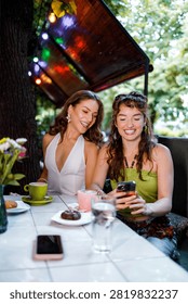Two friends sit at a cafe table outside, laughing and looking at a phone while enjoying drinks and snacks.