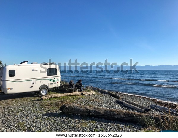 Two friends sit and\
read in lawn chairs after parking their holiday trailer at a scenic\
roadside rest stop along the Pacific coast line near Sooke, British\
Columbia, Canada 