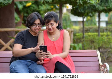 Two Friends Sitting On A Red Park Bench Looking Into The Mobile Phone And Smiling And Laughing In New Delhi, India