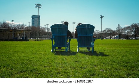 Two Friends Sitting On Wooden Chairs In The Park. Back View.