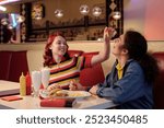 Two friends sitting in retro diner sharing a meal and milkshakes with playful gesture. Surroundings showcase a nostalgic ambiance with vibrant decor and classic diner elements