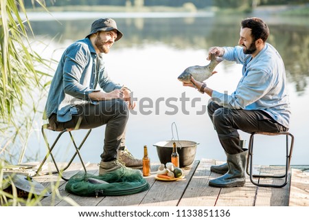 Two friends sitting together with beer and fish on the picnic while fishing near the lake