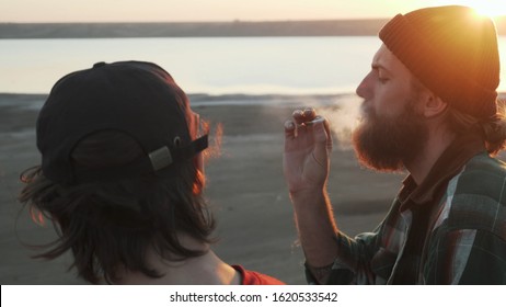 Two Friends Smoking Joint With Weed At Seashore At Sunset