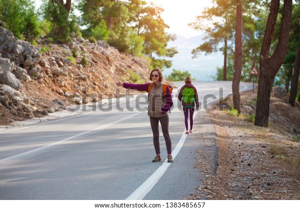 Two friends stop the car. Hitchhiking. Women\
catch the car on a mountain road. Girls with backpacks are on a\
mountain road. Free\
transport.