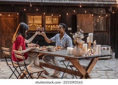 Two friends are toasting with glasses of red wine while sitting at a beautifully decorated table enjoying a delicious meal in a cozy outdoor setting - Powered by Shutterstock