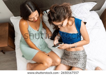 Two friends are using a smartphone while sitting on a bed, getting ready to go out and have fun