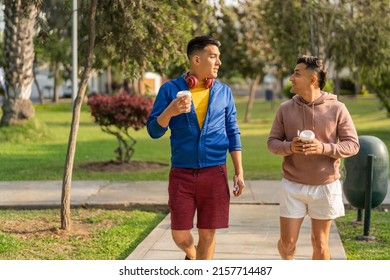 Two Friends Walking While Talking Along An Urban Park With A Take Away Coffee In A Sunny Day