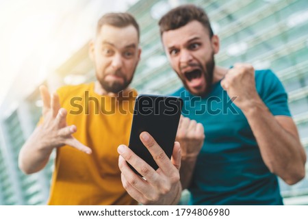 Two friends watching football play live broadcast online on mobile phone and cheering for favourite team with stadium on the background. Focus is on hand with phone.