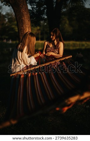 Two friends work together to set up a rainbow striped hammock in a park at golden hour. They share a quiet, warm moment outdoors, capturing friendship, leisure, and togetherness.