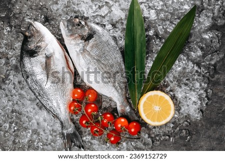 Two frozen dorado fish, which are located on ice particles on the kitchen table in one direction, next to which are two sprigs of a green plant, a branch with cherry tomatoes and a cut half of a lemon