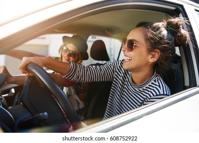 Two fun young women in sunglasses driving in a car in town laughing and smiling as they socialise together, view through open side window