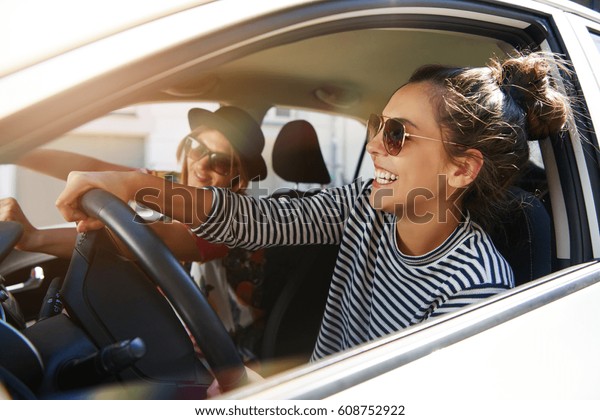 サングラスをかけた2人の楽しい若い女性が 街で車を運転しながら笑い 笑いながら付き合い 開いた窓から見る の写真素材 今すぐ編集
