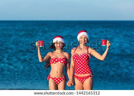 Two funny girls dressed in Christmas style and with cups in their hands. Summer Christmas celebration on sea beach