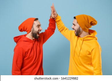 Two Funny Hipster Men Guys In Fashion Red Yellow Clothes Posing Isolated On Pastel Blue Background Studio Portrait. People Lifestyle Concept. Mock Up Copy Space. Giving High Five Holding Hands Folded