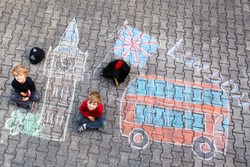 Two Funny Kid Boys In British Queen's Guard Soldier And Policeman Uniform Having Fun With London Picture Drawing With Colorful Chalks. With Big Ben, Union Jack And Red Bus.