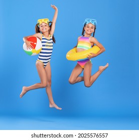 Two funny little girls sisters in swimsuits and swimming goggles jumping up in air with inflatable swimming ring and ball in hands on blue studio background, excited and ready for summer sea vacation