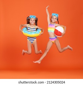 Two funny little girls sisters in swimsuits and swimming goggles jumping up in air with inflatable swimming ring and ball in hands on orange studio background, excited and ready for summer   vacation