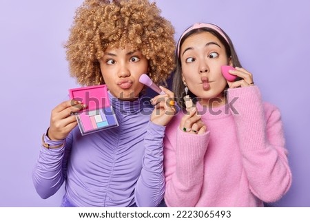 Two funny women do makeup apply eyeshadow and foundation make funny grimace fish lips prepare for party want to have fabulous look dressed casually isolated over purple background. Beauty procedures