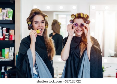 Two Funny Young Girlfriends In Hair Curlers Wearing Capes Having Fun Time Together In Beauty Salon. Female Friends Fooling Around With Rollers, Making Faces