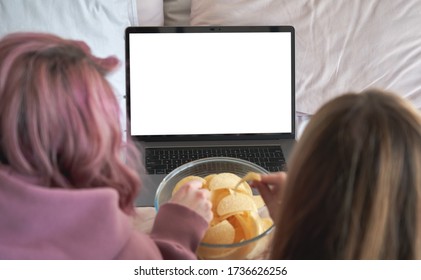 Two gen z teen girls friends watching tv video movie series, online reality show on laptop computer white mock up screen using digital streaming service subscription eat chips snack in bed. Back view - Powered by Shutterstock