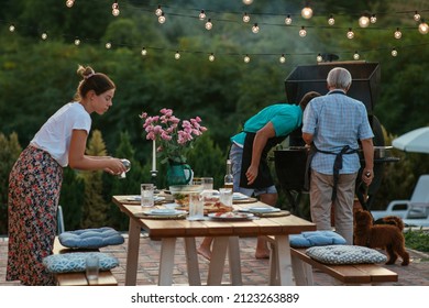 Two Generations Of Family Preparing Dinner. Young Adult Woman Setting The Dining Table And Male Members Barbecuing Beside Her.