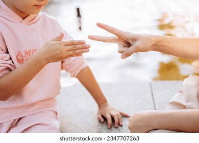 Two girl friends are playing a game of paper scissors. Caucasian children sitting by the fountain outdoors, playing together. An interesting and entertaining activity for children. - Powered by Shutterstock