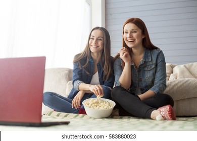 Two girl friends sitting on the floor near the couch in front of them there is a computer on which they watch a movie and eat popcorn. Funny friends relaxing together.