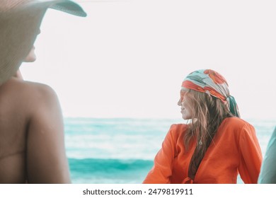 Two girlfriends are sitting on the beach during vacation. A red-haired woman in orange sunglasses smiles and looks away. The second woman in a hat sits with her back to the camera. Holiday with friend - Powered by Shutterstock