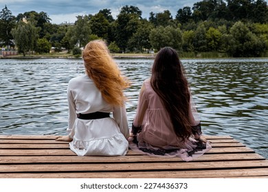 two girlfriends are sitting on the platform against the background of the river - Shutterstock ID 2274436373