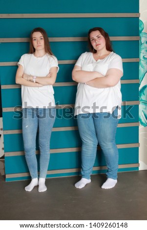 Two girlfriends, a slim blonde and a fat brunette, pose in a studio against a turquoise wall. Cute and different girls in blue jeans and white t-shirt