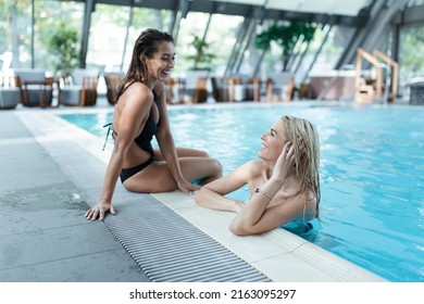 Two girlfriends in swimwear sitting on the poolside, enjoying summer vacation on the swimming pool intdoors - Powered by Shutterstock