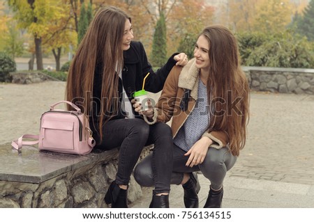 Two girlfriends try to warm up with a hot drink in the outdoors. They are having fun and fooling around, talking on the smartphone and sharing events with each other