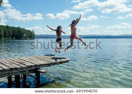Two girls in bathing suits jumping from a wooden pier into the water against a background of blue mountains in the sunlight