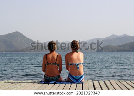 Two girls in bikini sitting on wooden pier on sea and misty mountains background. Beach vacation and travel, summer leisure and dreaming