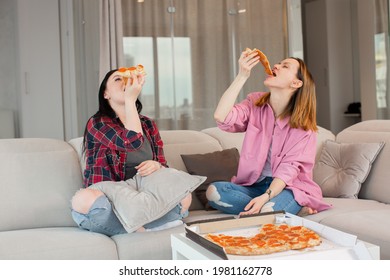 Two girls cheerfully put in their mouths pieces of pizza, laughing sitting at home on the beige sofa. High quality photo - Powered by Shutterstock