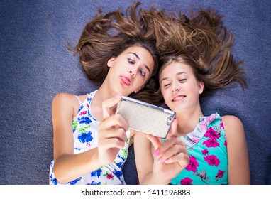 Two Girls In Colorful Summer Dresses Lying On The Bed Looking At Smart Phone, Taking Selfie For Social Media, Smiling, Being Goofy. Social Media Concept