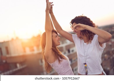 Two girls dancing on a building rooftop at sunset, affectionate and happy, with cityscape in the background - Powered by Shutterstock