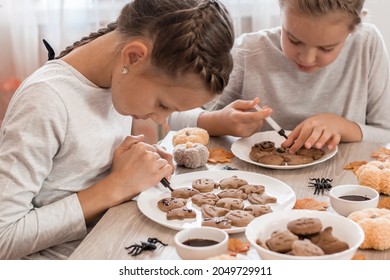 Two girls decorate halloween gingerbread cookies on plates with chocolate icing. Cooking treats for halloween celebration. Lifestyle