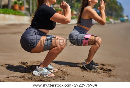 Two girls do fitness on the beach. They make squats with some ribbons. Booty Bands