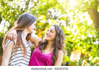 Two girls embraced together at park. They are happy and smiling while looking each other. Best friends embracing with trees on background. Lifestyle and friendship concepts.