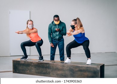 Two Girls Exercising With A Personal Trainer At The Gym With Protective Mask In Pandemic Period Of Covid19 - Two Fit Young Girls In A Gym Doing Squats With Their Afro American Trainer