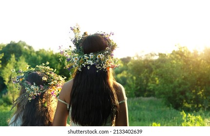 Two girls in flower wreaths on meadow, sunny green natural background. Floral crown, symbol of summer solstice. Slavic ceremony on Midsummer, wiccan Litha sabbat. pagan holiday Ivan Kupala