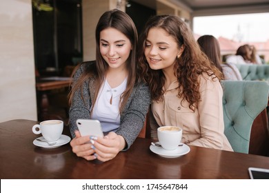 Zwei Mädchen Freund trinken Kaffee im Café und schauen in Smartphone. Herzlichen Glückwunsch an die Frauen, die auf der Terrasse sitzen. Menschen sehen Geschichten aus