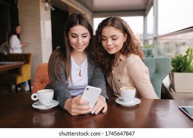 Zwei Mädchen Freund trinken Kaffee im Café und schauen in Smartphone. Herzlichen Glückwunsch an die Frauen, die auf der Terrasse sitzen. Menschen sehen Geschichten aus