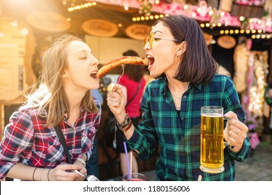 Two Girls Friends Having Fun And Eating Fried Sausage And Drinks Mug Of Beer At The Fair Market Square In Germany, Beer And Local Food Festival Concept