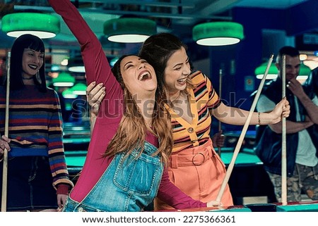 Two girls in a game room holding pool cues and cheering after making a successful shot, with other friends laughing in the background